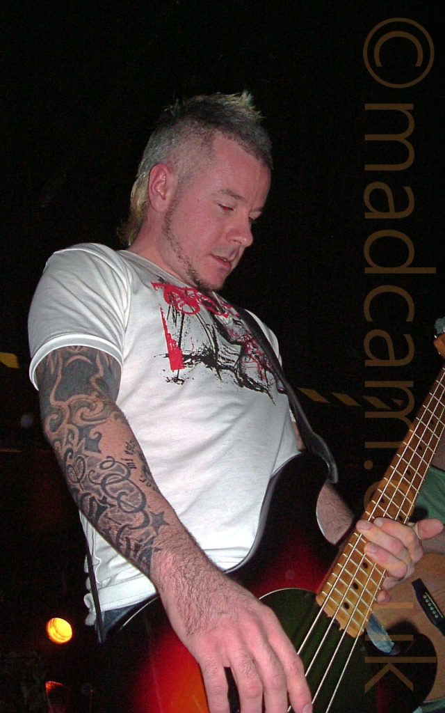 A man with short, blond, spiky hair, wearing a white t-shirt with a large black and red image across the chest, playing a red and black bass guitar. His right arm is covered in black tattoos. He appears to be looking at the floor in front of him.The background is mostly black, with a black and yellow stripe running along the wall behind him, and an orange light shining in the bottom right corner.