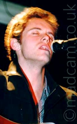 Long exposure shot of a young man with short dark hair and wearing a black coat over a blue shirt, singing into a microphone on a stand in front of him. The top of a red acoustic guitar can be seen at the bottom left of the frame. Yellow light from behind illuminates his hair, appearing a it frizzy as he moves his head during the exposure. A strip of red light behind the opening in his coat shows where his body has moved. The room is dark behind him.