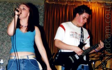 Young woman with long red-ish hair, wearing a blue crop-top and blue jeans, singing in to a microphone held in her right hand on the left of the frame, with a young man with short, black hair, roughly spiked, wearing a white t-shirt with red slashes over the shoulder and playing a black guitar, standing behind her on the right of the frame. Greeney-grey wallpaper covers the wall on the left, with golden curtains on the wall on the right.