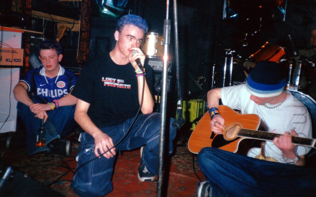 Several members of a band on stage - in the centre is a young man with short, dark hair that has been dyed blue, wearing a black t-shirt with red and blue text reading "Lost Prophets", and blue jeans. He is crouched down, and singing into a microphone held in his left hand. On the left of the frame is a young lad with short dark hair, wearing a purple t-shirt with a white collar and white text "Phillips" on the chest, and blue baggy jeans. He is sitting on the floor, his legs bent and crossed in front of him, holding a beer bottle and staring blankly at the floor. On the right of the frame is another young man, his head covered in a black, blue, and white banded woollen hat, wearing a white t-shirt and blue jeans, and playing an acoustic guitar. He is sitting on the floor with his les crossed beneath him. The stage is covered in an elaborately decorated red carpet. In the background, various pieces of on-stage equipment can be seen lining the black-painted walls - amplifiers, speakers, drums, crates, boxes, guitars.