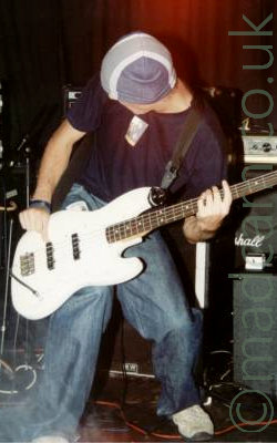 A man playing a large white bass guitar. He is slightly hunched over, and is wearing a grey and white hat, a black t-shirt with a white square logo in the middle of the chest, and blue baggy jeans, with white trainers. Behind him on the right are some amplifiers, while the drum kit is on the left. A red curtain acts as a backdrop, highlighted by a red light shining on the middle.