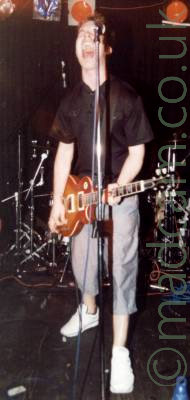 A man standing on a stage, screaming into a microphone held on a stand. He has short dark hair, and is wearing a black shirt and light grey trousers and white trainers, and is playing a brown electric guitar. In the background, a drum kit is in the middle of the stage, with black curtains behind that, with large coloured lights on roof-mounted racks at the top of the frame, pointing at the front of the stage.