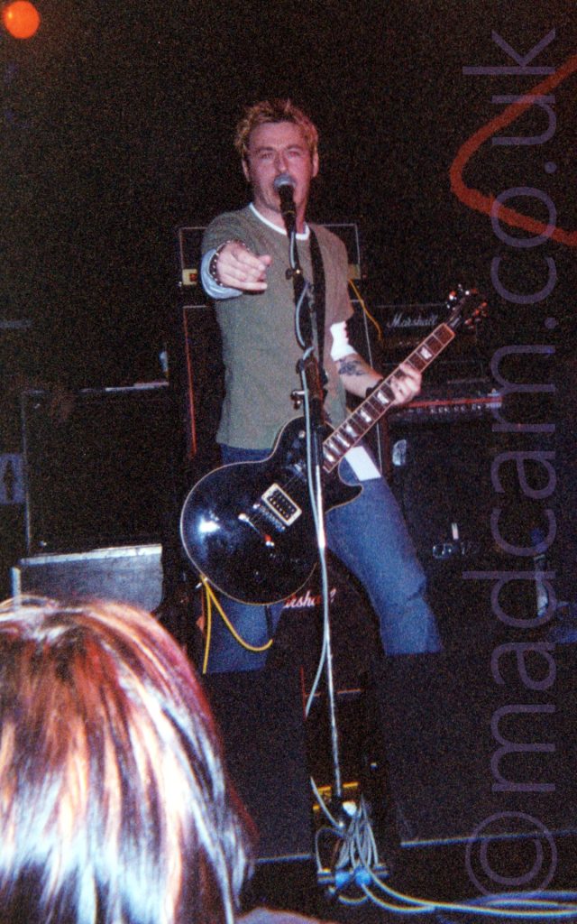 A man with short brown hair, wearing a long-sleeved white t-shirt under a green t-shirt, and a blue pair of jeans, holding a guitar, and pointing towards the camera. His mouth is hidden behind a microphone on a stand in front of him. The head of someone in the crowd is visible in the bottom left corner. A collection of amplifiers makes up a large part of the background, with black walls filling the rest.
