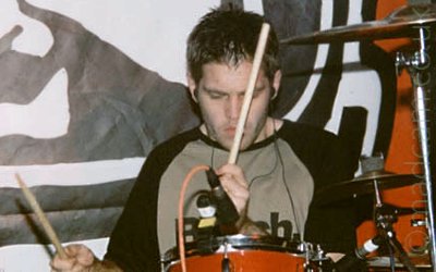A man with short dar hair and wearing a grey t-shirt with long black sleeves, sitting behind a red and chrome drum kit, holding a pair of drumsticks in his hands, staring at his drums with a look of concentration on his face. A microphone is mounted on the drum in front of him, with several cymbals on stands to his left, on the right of the frame. Behind him is a red logo with a black and white ink drawing of a face in a helmet.