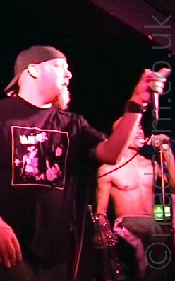 Vertically-oriented, low-resolution shot of a white man with a bushy goatee beard, and wearing a backwards baseball cap on his head, and a black t-shirt bearing an image of 2 people in a band, one of whom is playing a guitar, with some indistinct text at the top. He is holding a microphone in his left hand, and appears to be gesturing towards the crowd. Behind him, a bare-chested black man is singing into a microphone on a stand, holding the mic in his left hand, and a trumpet in his right. A green light in the background is shining down by his waist. Black walls and ceiling fill the rest of the frame The whole scene has a red tinge from the stage lights.