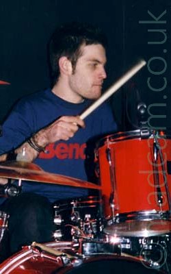 Close-up of a man with short dark hair and wearing a blue t-shirt with the word "Bench" emblazoned across the chest, sitting behind a black and red drum kit, holding a stick in his right hand, the stick starting to blur as he brings it down towards the drum in front of him. His right leg is visible behind the upright bass drum at the bottom of the frame. A golden cymbal cuts into frame from the left, between his leg and his arm. Black walls fill the background.