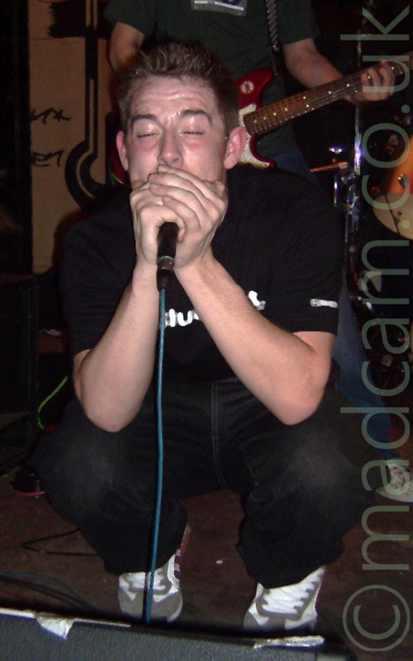 The singer from a band, crouching down on stage and singing into a microphone held in his hands, his eyes closed. He is wearing a black t-shirt and black jeans, and has short, spiky brown hair A man wearing a green t-shirt and playing a red and white guitar can be seen in the background