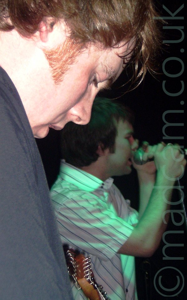 Closeup of the head and shoulder of a man with blond hair, looking down, taken from his right. He has sideburns reaching down to the base of his ears, and is wearing a grey shirt. The head of his guitar can be seen low down in the middle of the frame. Beyond him is a man with dark hair covering his ears, wearing a white shirt with thin black stripes, and singing in to a microhone on a stand in front of him, cupping the mic in both hands. Black walls fill the rest of the frame.
