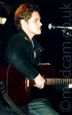 Side view of a man facing to the right, playing a dark brown acoustic guitar, wearing a black jacket over a grey shirt, and a dark pair of jeans. His short, dark, curly hair is being illuminated from the rear by an overhead red light. He is facing a microphone on a stand, which he is singing into, with his eyes closed. The background is almost entirely black, the colour of the walls in the poorly lit room.