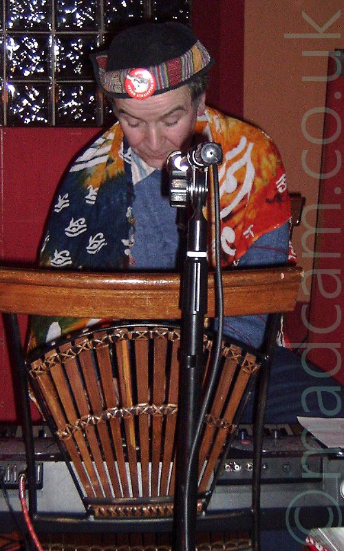 A seated man with dark grey hair covered by a black hat, wearing a grey jumper over a striped white shirt and blue trousers, his shoulders draped with a blue, orange, and yellow piece of cloth covered with white eye logo's. He is playing a keyboard that has been placed on a chair whose back is facing the audience. A microphone on a stand is in front of him, positioned over the top of the chair, as he looks down at his keyboard. An orange and red wall fills the background, with a square of glass bricks in the top left corner.