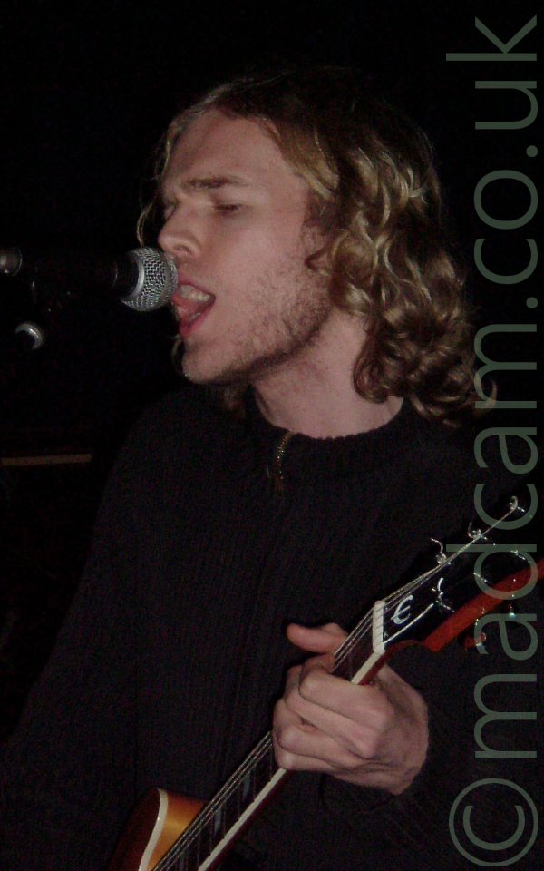 Head and shoulders shot of a man with long blond hair and stubble on his jaw, wearing a brown top, and singing into a microphone held on a stand in front of him, while playing an electric guitar. His eyes are almost completely closed. The walls in the background are almost entirely black.