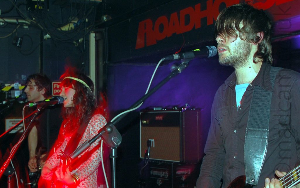 Three members of a band, lined up side-by-side at the front of a stage, all singing into microphones on stands in front of them, and playing guitars. On the left is a man with short, wavy brown hair and wearing a black t-shirt, in the middle is a woman wearing a flower print dress, her long, dark, wavy hair cascading over her shoulders, and with a gold-coloured band around her head. On the right is a man with unkempt, slightly long dark hair, covering his eyes at the front, and wearing a grey shirt over a blue t-shirt. In the background, the walls and ceiling are black, with red "Roadhouse" titles on a beam over a purple alcove.