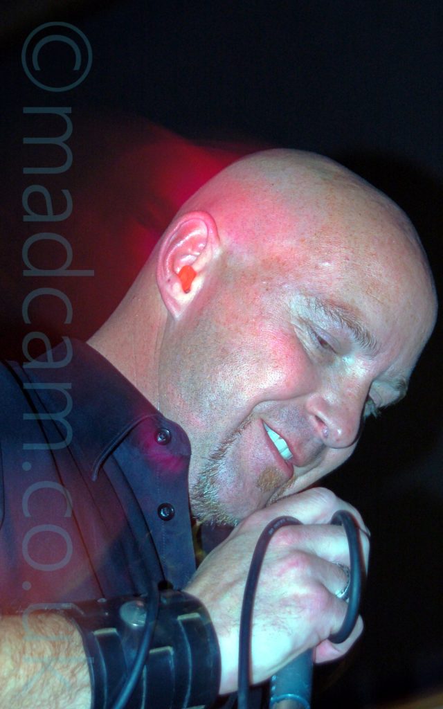 Close-up of the face of a shaven-headed man with a small goatee beard, wearing a black shirt, and holding a microphone in his right hand up towards his mouth. He has a slight smile on his face. A strong burst from a flash has frozen him in place, and the long exposure shows red streaks of light where he has moved his head backwards while the shutter was open. Black walls fill the rest of the frame.