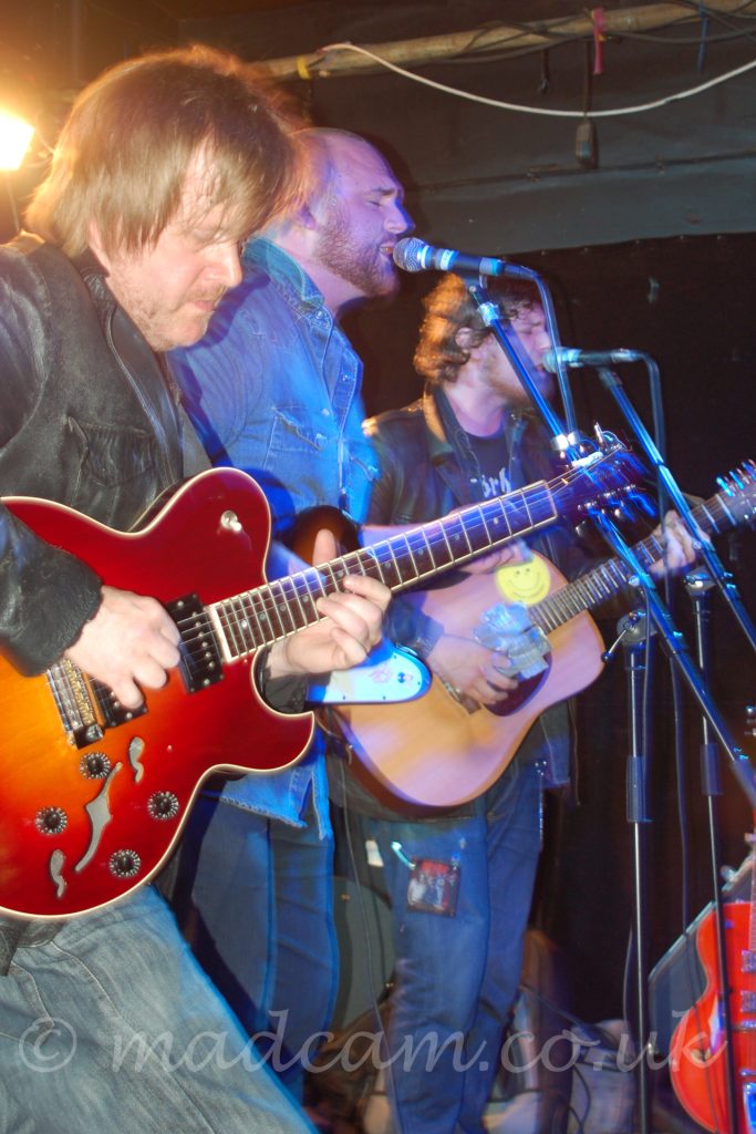 A view from the side of a stage. 3 men lined up side by side at the front of a stage, all playing guitars, the furthest 2 singing into microphones on stands in front of them. Nearest the camera, on the left, is a man with brown hair and beard, wearing a leather jacket and faded dark blue jeans, playing a red electric guitar. In the middle is a man with very close cropped dark hair and beard, wearing a denim shirt and pants, , playing a brown and white electric guitar, his eyes closed as he sings. Last is a man with curly brown hair, wearing a leather jacket over an open denim shirt over a black t-shirt, as well as dark blue jeans, playing a brown acoustic guitar and singing, with his eyes shut as well. In the bottom right corner of the frame, a red electric guitar sits against a monitor speaker. The walls are black, with dirty grey ceilings, and a light at the back of the stage shining almost directly into the camera. There is some movement in the frame, from the slightly longer-than-usual exposure used.