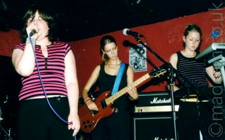 3 women in a band - singer, on the left, with short dark hair and a pink and black striped top, singing into a microphone. Guitarist in the middle, with long tied back blonde hair, wearing a black tank top and playing a brown electric guitar. Keyboard player, with blonde hair tied back, wearing a black and grey top, and playing 2 banks of keyboards stacked vertically. Ruby red walls and black ceiling fill the background.