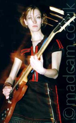 Long exposure shot of a woman wearing a black top and skirt with red highlights, playing an electric guitar. A flash has been used to freeze her in space, and swirls of red light show where she has moved afterwards. Discrete points of light in the darkness behind her head have turned into white streaks.