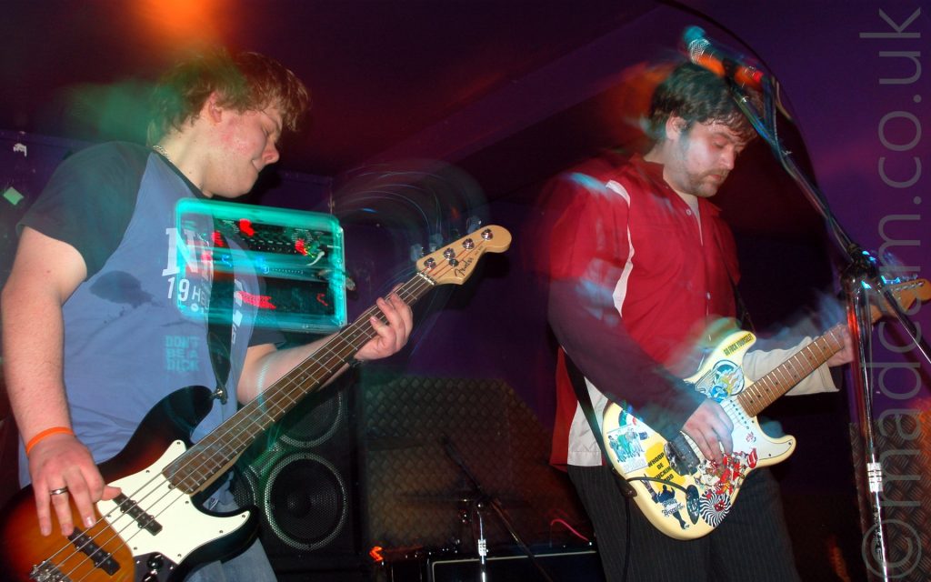 Two members of a rock band playing their instruments. On the left is a man with brown hair, wearing a blue t-shirt with dark blue sleeves and almost indecipherable white writing on the chest, and playing a 4 stringed bass guitar. Behind him is a large amplifier with 4 speaker cones, and 2 glowing 2 racks on the top. The racks seem to shine through his body. Ion the right is a wan with dark hair and a bit of facial stubble, wearing a red and white shirt and dark grey pin-stripe trousers, playing a yellow guitar adorned withlots of stickers. A microphone on a stand is in front of him. The walls and ceiling behind them are purple. The photo is taken with a long exposure time, meaning that as both men move, they and their instruments leave light trails.