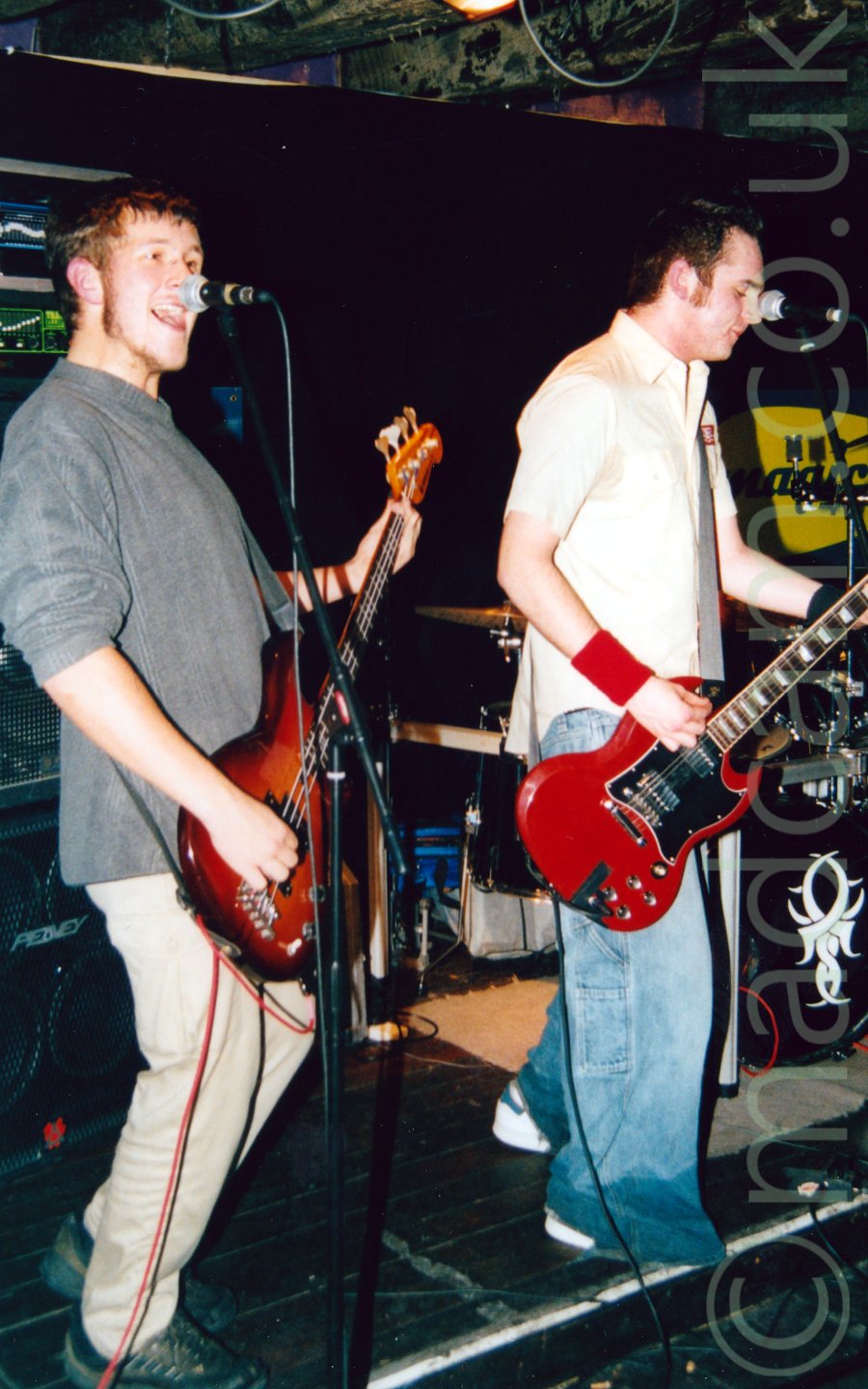 2 musicians performing on a stage. On the left is a man with short brown hair and a beard that runs along his jawline, wearing a grey jumper and light coloured trousers. He is playing a bass guitar and singing into a microphone held on a stand in front of him. On the right is a man with spiky dark hair, wearing a light-coloured shirt and blue baggy jeans, and holding a red and black electric guitar. A tall, black amplifier is visible in the background on the left, with a drumkit visible ion the right. Black walls and a partial black ceiling fill the rest of the frame, with exposed beams right at the top.