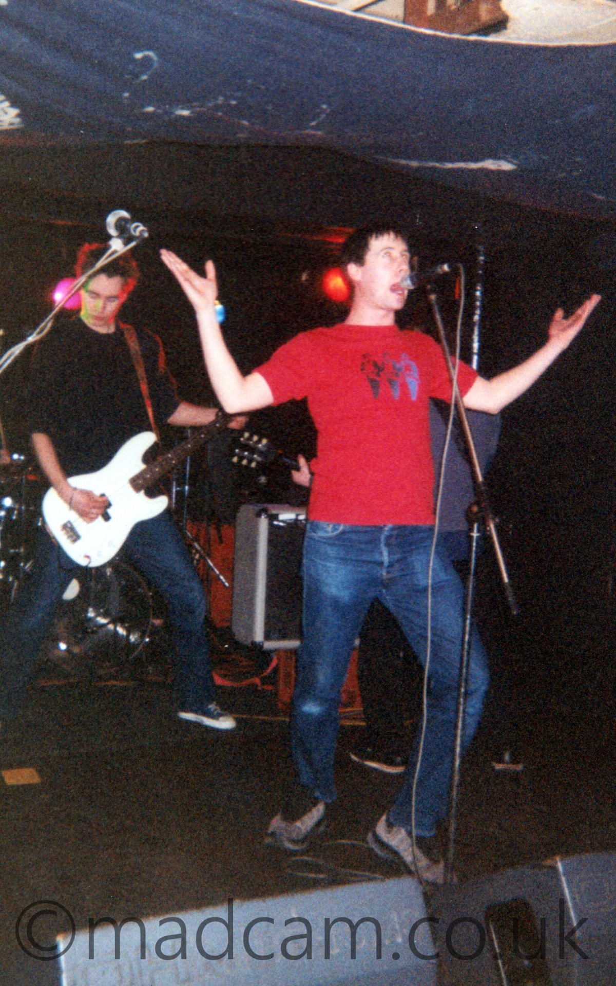A band performing on a stage. In the foreground is a man with short dark hair, wearing a red t-shirt and blue jeans, singing into a microphone on a stand in front of him. His knees are bent, almost crouching, while his arms are spread wide, his hands level with his head. Behind him, on the left of the frame, a man with dark hair and wearing a black jumper and blue jeans is playing a white guitar, looking down at the ground. On the singers other side, a man in a grey shirt is standing behind him, his back turned to the audience. A white amp is in the middle of the frame in the background. The rooms walls are black, while the white roof has a black tarp covering the back section.