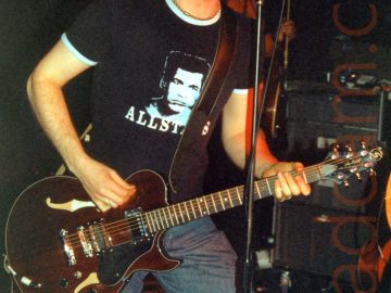 A man with short brown hair, wearing a dark blue t-shirt with a photo of Muhammed Ali on the chest and the word "Allstars" underneath, and a pair of blue jeans. He is playing a brown guitar, and singing into a microphone on a stand in front of him. In the background, a man in a red shirt is holding a guitar. Black walls and ceiling fill the rest of the frame.
