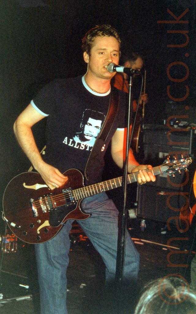A man with short brown hair, wearing a dark blue t-shirt with a photo of Muhammed Ali on the chest and the word "Allstars" underneath, and a pair of blue jeans. He is playing a brown guitar, and singing into a microphone on a stand in front of him. In the background, a man in a red shirt is holding a guitar. Black walls and ceiling fill the rest of the frame.