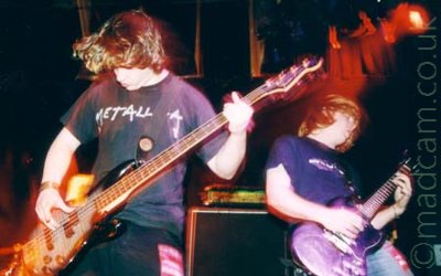 2 members of a band performing on stage-on the left, a man with brown hair, wearing a black t-shirt and dark blue jeans, playing a black and blue bass guitar, leaning forwards, with another man on the right, with unkempt ginger hair, and wearing a dark blue t-shirt and light blue jeans, playing a purple electric guitar and leaning backwards. Both men are frozen in place by the flash, but are still obviously moving, leaving coloured shadows at the edges of their arms and heads. A red light in the upper right corner is painting the center of the frame, creating visual echoes of the head of the bass. Black walls with mounted wooden frames fill the background.