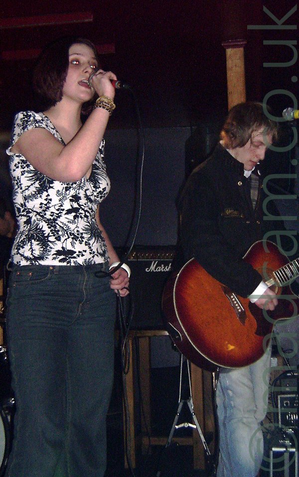 2 members of a band performing on stage, a woman with short brown hair and wearing a white top with a black flower motif and dark jeans, holding a microphone that she is singing into, and a man with short, light brown hair and wearing a dark jacket and pale blue jeans, playing a brown acoustic guitar. Dark walls and ceiling fill the background, with a black amplifier on a stool in the middle of the frame.