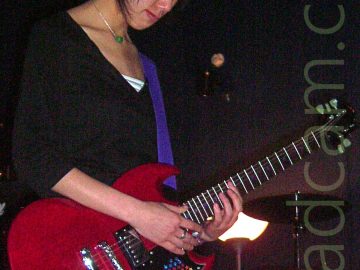 A woman with long black hair tied back with a purple scrunchie, wearing a black top and grey-blue jeans, playing a red electric guitar while gazing at the floor. The guitar is attached to a purple strap slung over her left shoulder. Black walls and ceiling fill the background, with a white light shining in the middle of the frame.