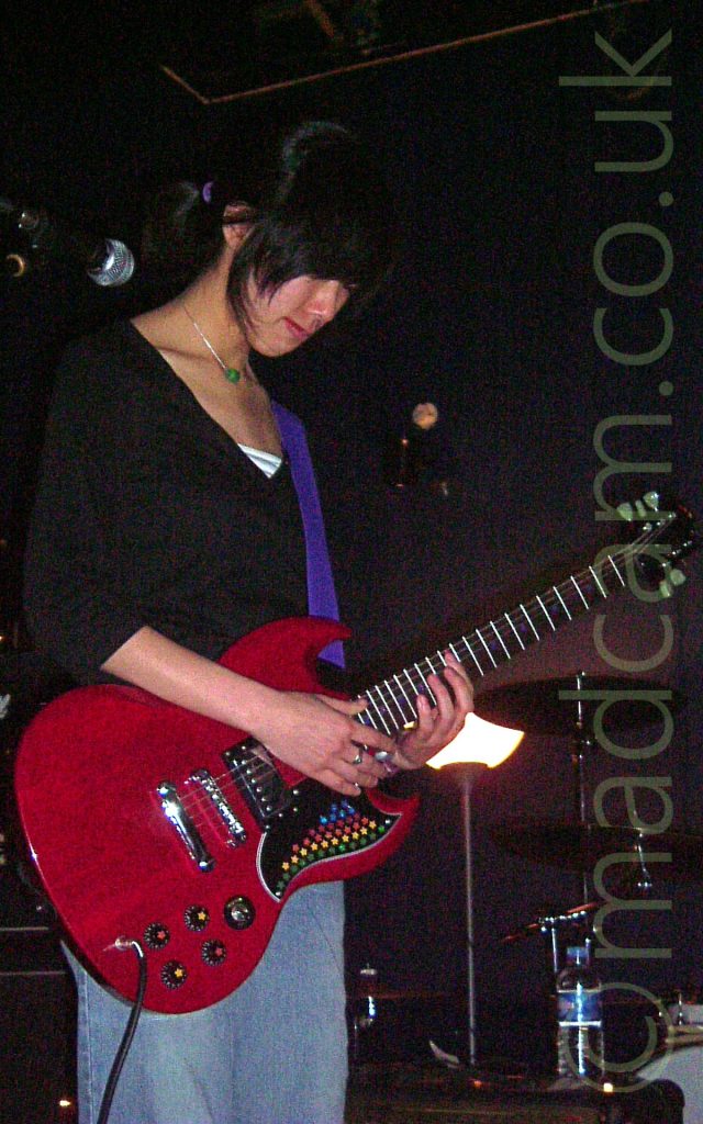 A woman with long black hair tied back with a purple scrunchie, wearing a black top and grey-blue jeans, playing a red electric guitar while gazing at the floor. The guitar is attached to a purple strap slung over her left shoulder. Black walls and ceiling fill the background, with a white light shining in the middle of the frame.