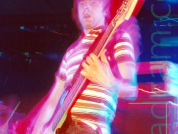 A man with short, dark hair swept down over his forehead, wearing a whit polo shirt with brown and orange bands around the body, and a blue pair of jeans, standing on a stage and playing a brown and white bass guitar. A flash has frozen him in time, while a long exposure, a slight zoom, his movement, and the stage lights have combined to paint the area around him in red. Amplifiers sit on the stage behind him, also tinted red, while the walls and ceiling at the back of the stage are black, with green speckles of reflected light on metal frames right at the back.