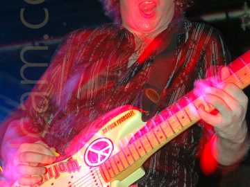 Long exposure image of a man with long, wavy brown hair, wearing a dark shirt with white vertical stripes and grey trousers, playing a yellow electric guitar, with his mouth open and eyes almost closed in almost ecstacy. The guitar is covered in various coloured stickers, with Pearl Jam and Rolling Stones band logos, a CND logo, and others. A flash has frozen him in place, and a long exposure creates movement, with streaks of red, blue, and purple light, and secondary images Black walls and ceiling fill the background, with a metal pole stretching across the top of the room behind his head.