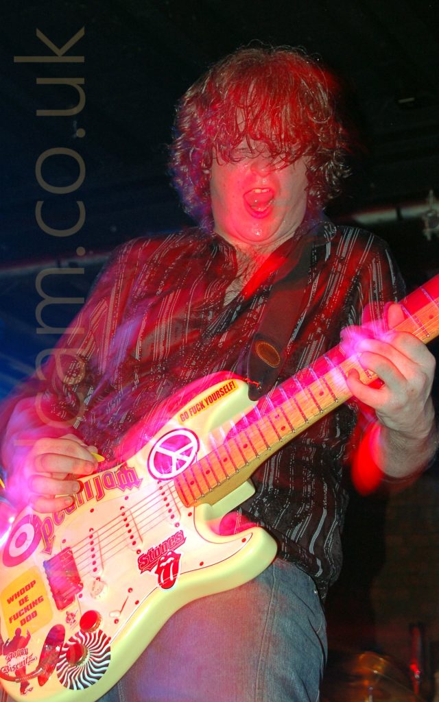 Long exposure image of a man with long, wavy brown hair, wearing a dark shirt with white vertical stripes and grey trousers, playing a yellow electric guitar, with his mouth open and eyes almost closed in almost ecstacy. The guitar is covered in various coloured stickers, with Pearl Jam and Rolling Stones band logos, a CND logo, and others. A flash has frozen him in place, and a long exposure creates movement, with streaks of red, blue, and purple light, and secondary images Black walls and ceiling fill the background, with a metal pole stretching across the top of the room behind his head.