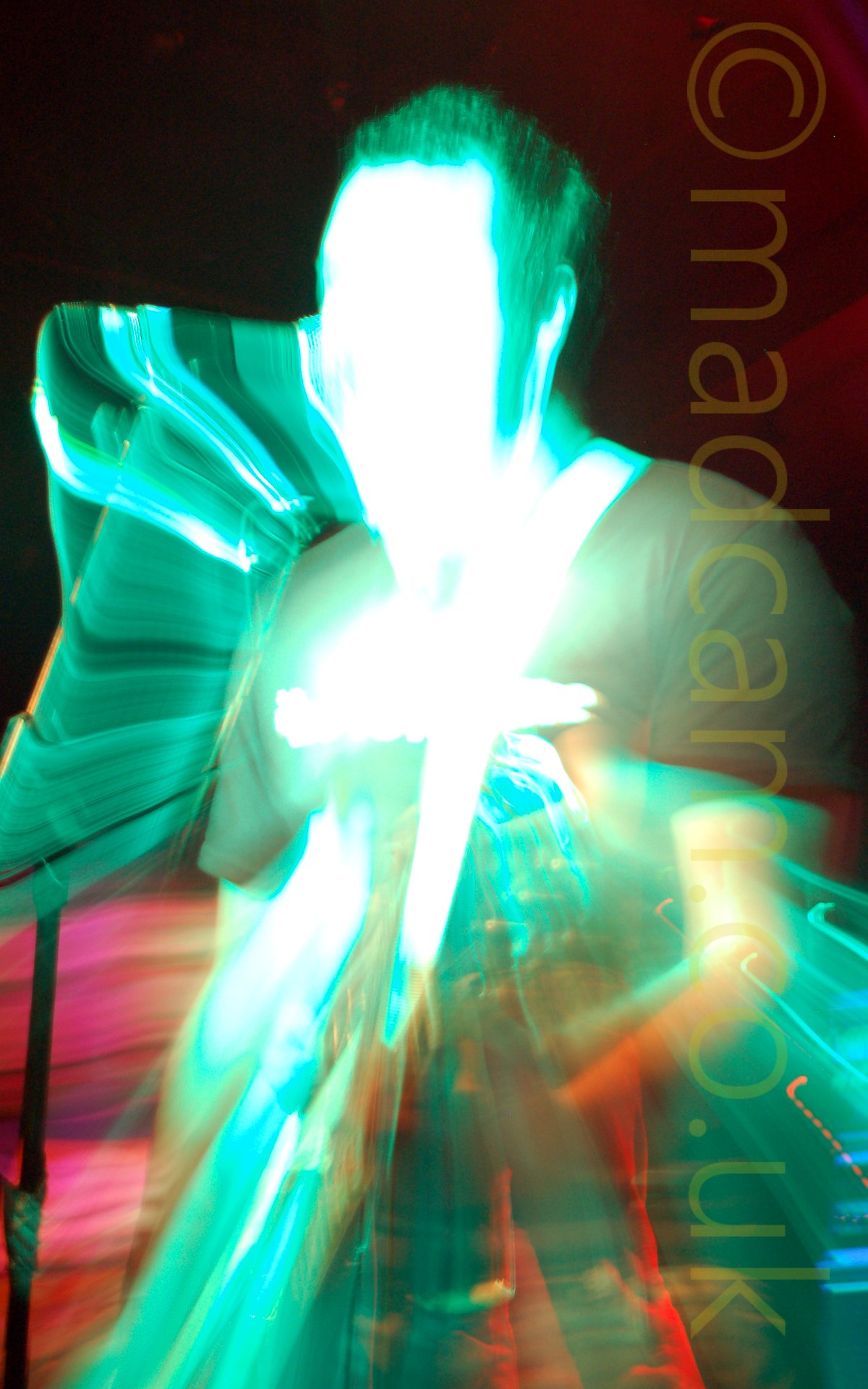 A rather colourful long exposure shot with a snap zoom of a man playing a guitar and singing into a microphone. One of the stage lights has washed out all details of his face, and both he and the microphone on it's stand appear to be shrinking and vanishing in to the middle of the frame, surrounded by green smears of light. Red walls and ceiling fill the rest of the frame.