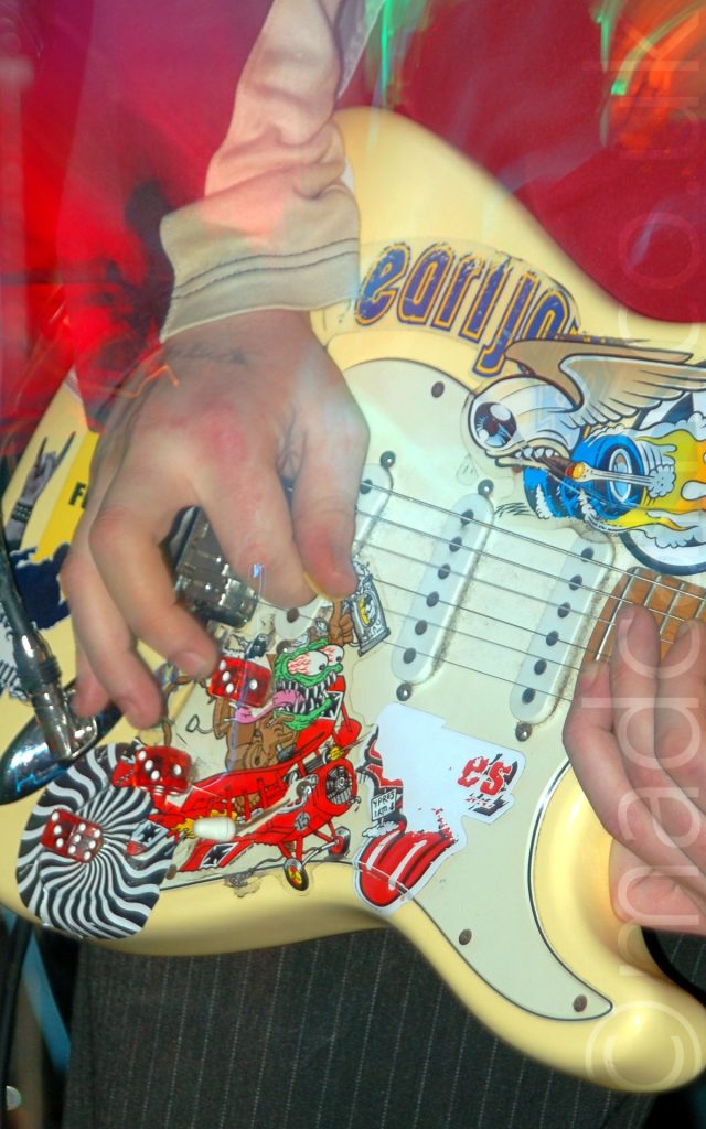 Close-up of the pale yellow body of a guitar being held by a man wearing a cream-coloured long-sleeved t-shirt and grey pin-striped trousers. There are a number of illustrations on the body of the guitar, including a red biplane, the red tongue from the Rolling Stones logo, a circular optical illusion, 3 red dice with white dots, and a winged eyeball smoking a cigar and rolling on thick wheels. The top of the image is painted in red light with green and orange streaks.