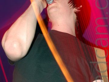 A man with dark hair and wearing a black t-shirt, holding a sunburst and white guitar in his left hand and a microphone on a stand in his right hand. He is singing into the mic while staring off into the distance. A long exposure has been used, a flash freezing him in the frame, and streaks of red light showing movement. Purple walls and ceiling fill the rest of the frame.