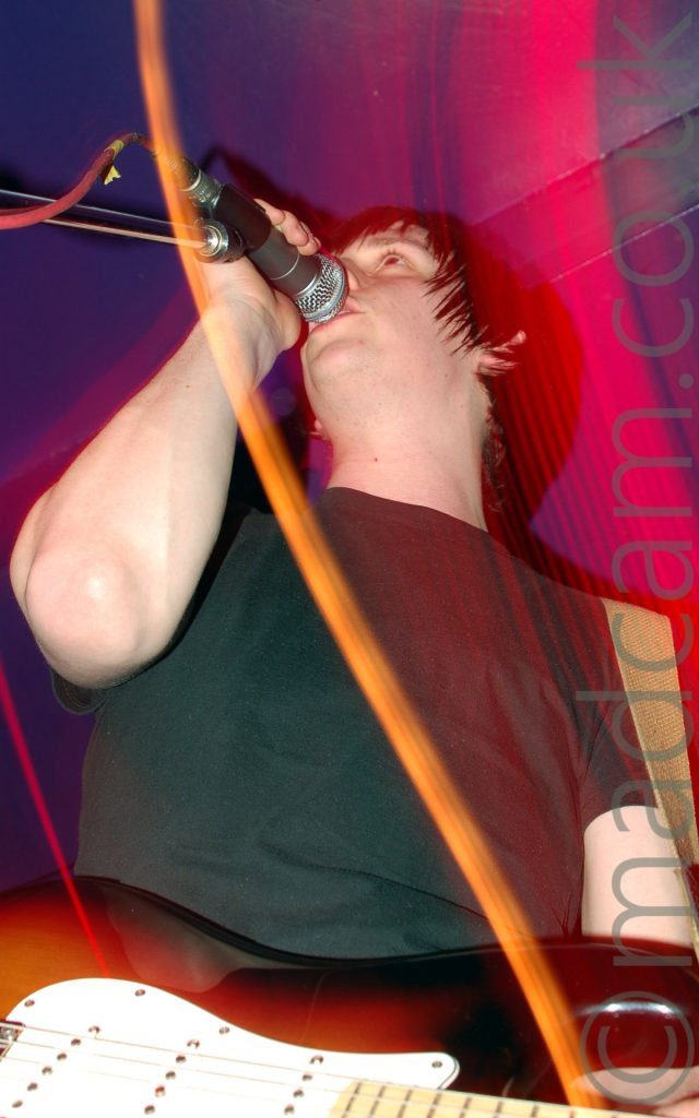 A man with dark hair and wearing a black t-shirt, holding a sunburst and white guitar in his left hand and a microphone on a stand in his right hand. He is singing into the mic while staring off into the distance. A long exposure has been used, a flash freezing him in the frame, and streaks of red light showing movement. Purple walls and ceiling fill the rest of the frame.