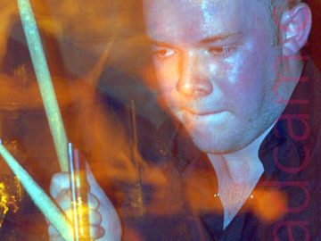 Close-up of a man with short, slightly spikey hair, wearing a black shirt, holding a pair of drumsticks raise in the air. A camera flash has frozen him in place, while a long exposure paints the frame with yellow lights.