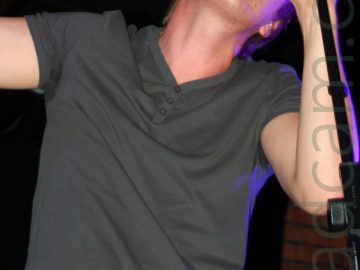 A man with short brown hair, wearing a green t-shirt and black jeans, leaning in to a microphone held on a stand in front of him, crooning in to the mic. Purple light is shining on his left side. Brown brick walls partially covered by black curtains are in the background, with black ceilings with ornate brown wooden roof supports filling the rest of the image.