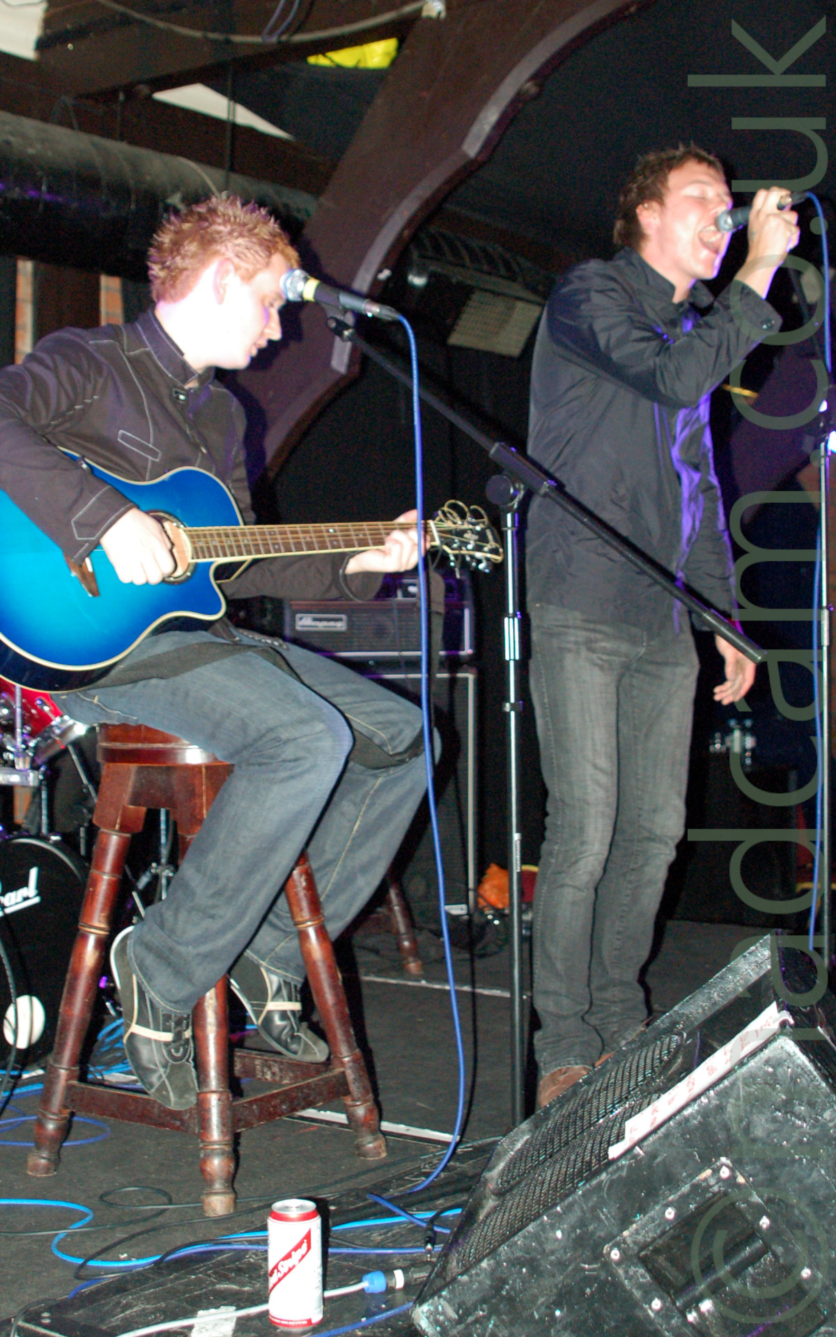 Vertically-oriented photo of 2 members of a band performing on a stage. On the left is a man with spiky ginger hair, wearing a black jacket and grey jeans, sitting on a tall wooden stool and playing a blue acoustic guitar. On the right is a man with short brown hair, wearing a black jacket and black jeans, standing behind a microphone stand which he is holding on to and singing in to. In the foreground, an angular speaker is on the floor, pointing more or less towards the man with the guitar, on the right of the frame, with a white and red beer can on the floor on the left. In the background, decorated brown wooden roof beams stretch across the frame, holding up the black ceiling.