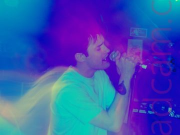 Long exposure of a man with dark hair, wearing a white t-shirt and a blue pair of jeans, holding a microphone in both hands as he sings. A long exposure shot, starting zoomed out then zooming in, with no flash. The result has clouds of blue and purple lights around his head, with streaks of green lights in both top corners, with secondary images of the singer behind him.