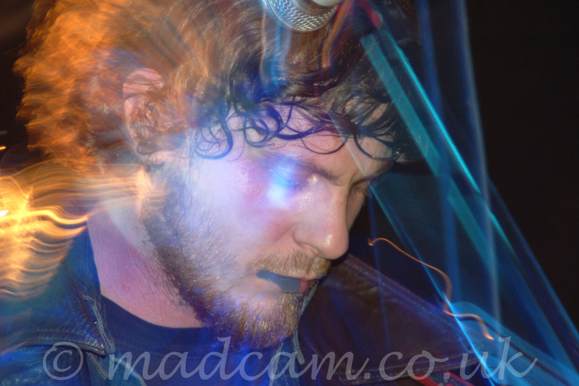 Long exposure close-up of the head of a main with wavy brown hair and a beard, wearing a black jacket over a black t-shirt, and holding a guitar pick in his mouth. This photo started off zoomed in close, and snap-zooms out during the exposure, leaving trails of blue and yellow coloured light, with the head frozen in place by an initial flash.