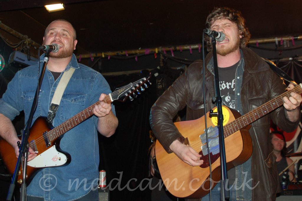 Two men performing on a stage. On the left is a man with Close-cropped hair and beard, wearing a blue denim shirt over a black t-shirt, and playing a brown and white electric guitar, and singing into a microphone on a stand in front of him. On the right is a man with long-ish, brown, curly hair, wearing a brown leather jacket over an open, black denim shirt over a black t-shirt with a white logo, playing a brown acoustic guitar, while singing (nay, screaming!) into a mic on a stand. A black curtain hides the walls in the background, with a metal frame lining the corners of the black ceiling.