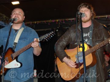 Two men performing on a stage. On the left is a man with Close-cropped hair and beard, wearing a blue denim shirt over a black t-shirt, and playing a brown and white electric guitar, and singing into a microphone on a stand in front of him. On the right is a man with long-ish, brown, curly hair, wearing a brown leather jacket over an open, black denim shirt over a black t-shirt with a white logo, playing a brown acoustic guitar, while singing (nay, screaming!) into a mic on a stand. A black curtain hides the walls in the background, with a metal frame lining the corners of the black ceiling.
