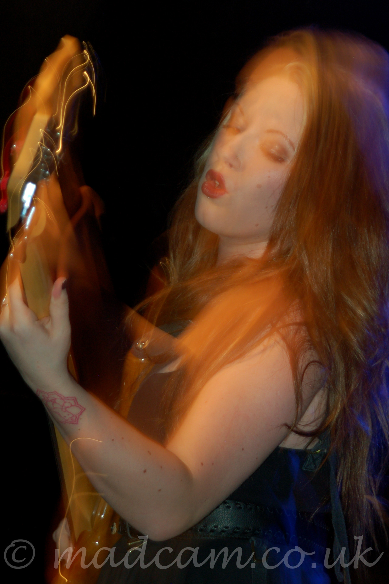 A long exposure close-up photo from the side of a woman with long blonde hair cascading over her shoulders, wearing a black dress that leaves her arms and shoulders bare, playing a brown bass guitar. She is leaning slightly backwards, and is looking at her left hand. There is a red tattoo on the back of her left wrist. A flash has frozen her in place, with streaks of lights showing how she has moved afterwards. The background is entirely black.