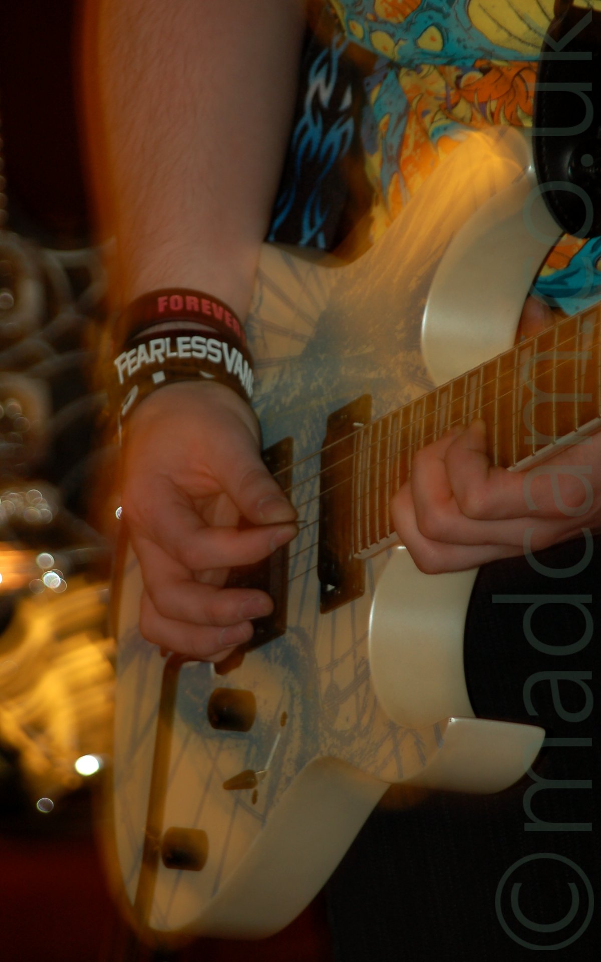Close-up of a white electric guitar being played by someone wearing a black t-shirt with a yellow and blue design on the front, and black pants, with several wristbands on their right wrist. Their right arm is frozen in place by the flash, but is also obviously moving, the long exposure painting it's edges with light. The background is blurred into smears of light.