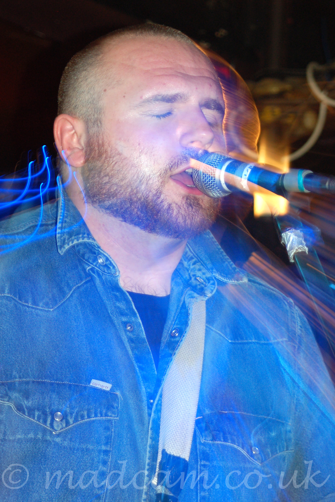Close up side view of a man with a short beard and cropped hair, with eyes closed, wearing a blue denim shirt, singing in to a microphone on a stand. This is a long exposure photos, and he is obviously moving, shadows and light streaks following him around the image.