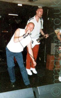 2 members of a band on stage - at the front is a shaven-headed man wearing a white t-shirt and blue jeans, holding a microphone in his right hand and singing into it, his eyes closed. The mic cord is wrapped around his wrist, and he is slightly bent over. Behind him is a man, also shaven-headed, wearing a white t-shirt and red trousers, playing a white bass guitar, and shouting into a microphone a couple of feet in front of him, with his eyes closed. He appears to be bouncing into the air. Black walls and a white ceiling fill the background.