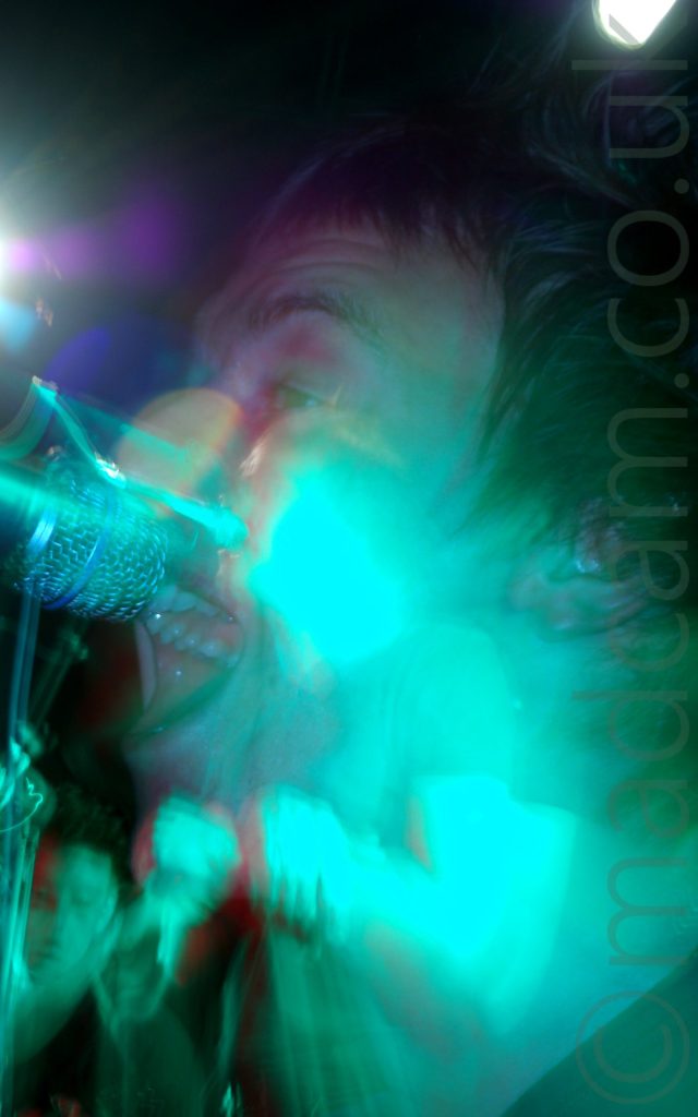 Close-up side view of the head of a man with dark hair, singing in to a microphone in front of his face. A long exposure has been used, along with a snap zoom out, so although his face fills the frame, there are secondary images around the edges of some of the other members of the band. The whole frame has a green tinge from the stage lights, with streaks of green, purple, and blue light throughout.