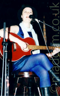 A woman wearing a black hat and a blue dress over blue leggings and black boots, sitting on a wooden brown chair and playing a red acoustic guitar. There is a microphone on a stand in front of her, which she is singing in to. Black curtains line the walls in the background, with a small section of brown brick visible through a gap.