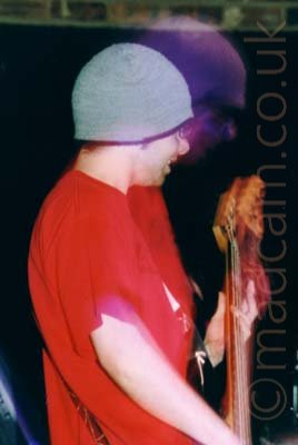 A man wearing a woollen grey cap and a red t-shirt, playing a long-necked bass guitar, standing facing to the right. He has been frozen in place by a flash from the camera, while a snap zoom creates a purple secondary image behind his head. The head of the bass is bathed in golden light. Black walls fill the rest of the frame.